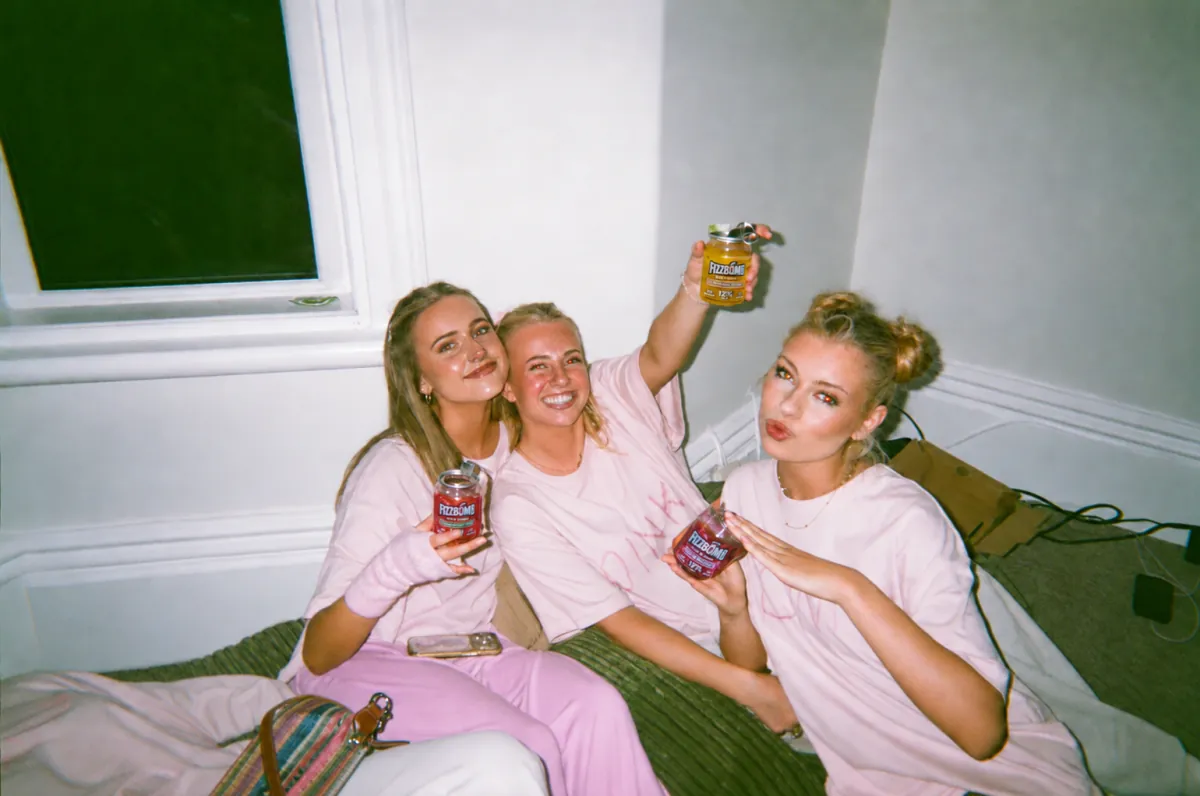 Three friends on a bed raising their FizzBomb cans