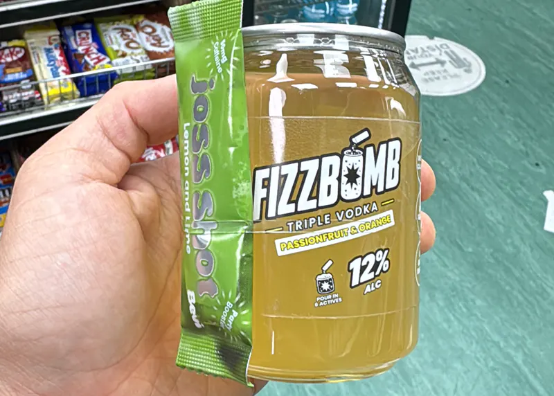 A FizzBomb Passion Fruit & Orange can held up in a corner shop, its sachet strapped to the side